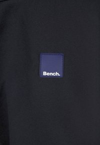 Czarny bawełniany t-shirt z prostokątną niebieską metką z napisem „Bench.” w białym kolorze. Gładka faktura z minimalistycznymi elementami designu.