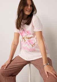 Šviesiai rožiniai marškinėliai su rožinės ir rudos spalvos gėlių raštu, ant kurio užrašyta „meilė čia auga“. Modelis dėvi rudas langeliais puoštas platėjančias kelnes.