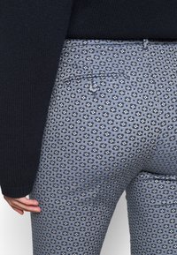 Pantaloni in denim con un motivo floreale blu e bianco, caratterizzati da un design aderente e un dettaglio della tasca sul retro, abbinati a un maglione scuro.