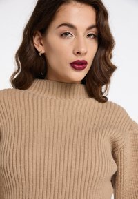 Maglione beige a coste con un collo alto e arrotondato. Tessuto morbido, presenta maniche lunghe e una silhouette aderente.