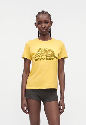 Kvinde med flettet hår bærer gul T-shirt med sovende kat og teksten "måske senere," kombineret med mørkegrå shorts, stående mod ensfarvet baggrund.
