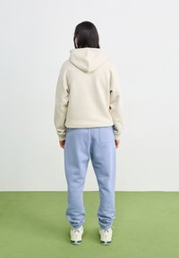 Hanorac bej cu glugă și pantaloni de trening bleu deschis, realizați dintr-un material moale. Pantalonii au o bicicletă pe spate. Încălțămintea este albă cu accente verzi.