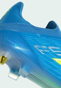 Chaussure de football bleue avec des crampons dorés et transparents, une tige en tissu texturé et des lettres blanches partielles sur le côté.