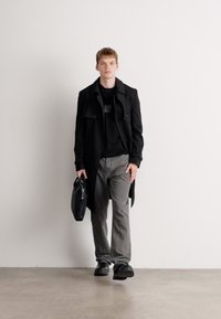 Black trench coat over a black shirt, grey trousers, and black shoes; holding a black bag; neutral background.