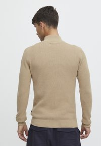 Beige strikket genser med en teksturert overflate, med lange ermer og en ribbestrikket halslinning. Bakvisning viser en tettsittende silhuett.