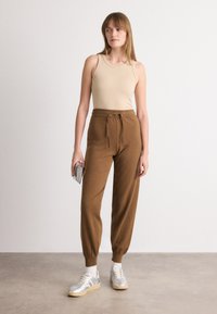 Top in beige abbinato a pantaloni jogger marroni con cordino. I jogger presentano polsini a costine e una consistenza morbida. La modella tiene una clutch argento, indossando sneakers argentate.