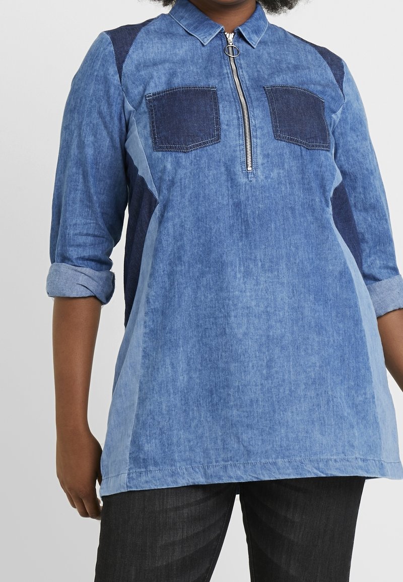 Tunic in denim di blu chiaro e scuro con chiusura a zip frontale, due tasche sul petto e maniche arrotolate, caratterizzata da una vestibilità rilassata e una texture morbida.
