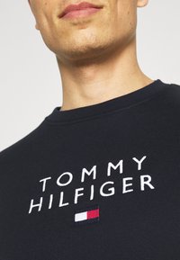 Granatowy sweatshirt z białym haftowanym napisem "TOMMY HILFIGER" oraz małym akcentem w kolorach czerwonym, białym i niebieskim poniżej napisu.