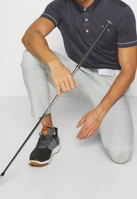 Pánská šedá polo košile s bílými detaily; světle šedé kalhoty; černo-šedé tenisky s oranžovým prvkem. Drží golfovou hůl.