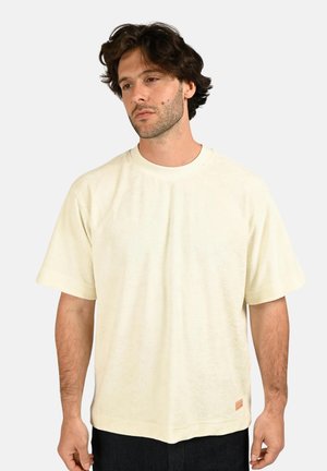 Homme aux cheveux foncés et ondulés portant un T-shirt ample écru à manches courtes et un pantalon foncé, debout devant un fond clair uni.