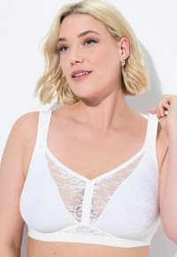 Witte kanten bralette met een V-vormig kanten paneel in het midden en brede banden. De stof heeft een zachte textuur en een bloemenpatroon.