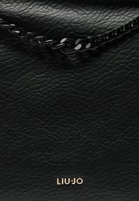 Sac en cuir noir avec une surface texturée, une bandoulière en chaîne métallique contrastante et un détail en or au centre en bas.