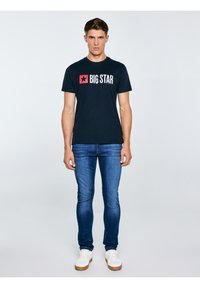 Mladić stoji, nosi crnu majicu s natpisom "BIG STAR", plave traperice i bijele tenisice na jednobojnoj svijetloj pozadini.