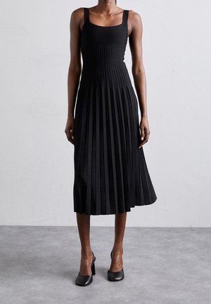 Femme portant une robe midi noire plissée sans manches et des chaussures slingback à talons noirs, debout sur un sol gris.