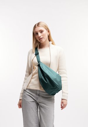 Jonge vrouw draagt een beige trui en grijze broek met een blauwe crossbodytas tegen een effen witte achtergrond.