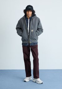 Grå hættetrøje med lynlås, ternet skjorte under, maroon bukser og hvide sneakers. Sort kasket med logo; krøllet hår. Struktureret stof.