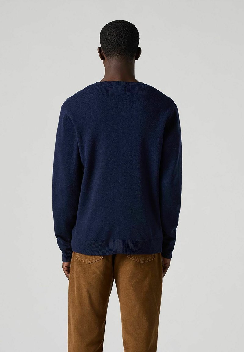 Dunkelblauer Strickpullover mit rundem Ausschnitt und gerippten Bündchen, getragen, kombiniert mit hellbraunen Cordhosen. Weiche Textur, lässiger Schnitt.