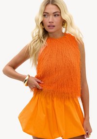 Femme blonde portant un haut sans manches orange texturé et une jupe assortie, accessoirisée avec de grandes boucles d'oreilles en or, un bracelet et des bagues.