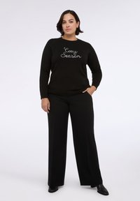 Maglione nero con la scritta "Cozy Season" in corsivo bianco sul petto, abbinato a pantaloni neri a gamba larga e scarpe nere, texture liscia.