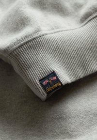 Sweatshirt gris clair avec des poignets côtelés, ornée d'un patch logo bleu marine avec des drapeaux croisés et le texte doré "Superdry". Finition douce et texturée.