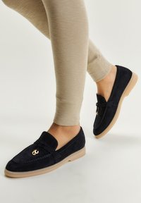 Mocasini din suede bleumarin cu talpă din cauciuc bej, cu un accent metalic auriu și cusături tradiționale de mocasin.