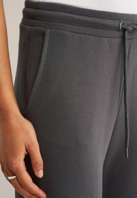 Sive trenirke od mekanog materijala imaju elastičnu traku s vezicama u struku i bočne džepove. Ruka je položena na tkaninu, pokazujući glatku teksturu.