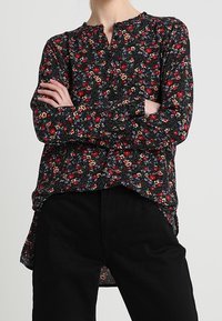 Svart blommig blus med långa ärmar, som har röda och vita blommönster, en rundad halsringning och en något längre baklinje.