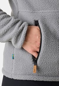 Grijze fleece jas met een gestructureerde oppervlakte, voorzien van een zwarte ritszak en een accent van de Zweedse vlag aan de zijkant.
