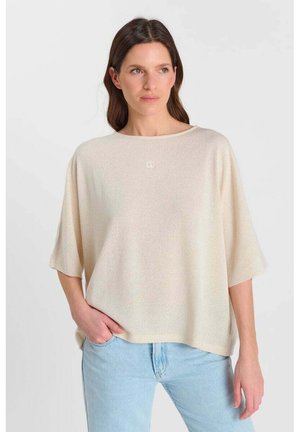 Vrouw die een loszittende, beige trui met korte mouwen draagt en een lichtblauwe spijkerbroek, staand met één hand in haar zak tegen een witte achtergrond.