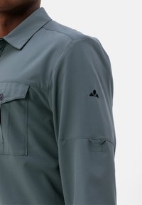 Camicia a maniche lunghe da uomo in teal smorzato, con colletto a bottoni, tasca sul petto e una trama tessuta. Accento del logo nero sulla manica.