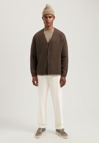 Bruine cardigan met zwarte knopen, gecombineerd met een beige shirt, witte broek en grijze sneakers. Model draagt een lichtbruine beanie.