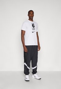 T-shirt branca da Nike com o logo da NBA, combinada com calças de treino pretas com detalhes em branco e pormenores em malha, usadas com ténis atléticos brancos.