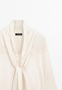 Blouse de couleur crème avec un col drapé noué en un nœud lâche, présentant un tissu subtilement texturé et une étiquette de marque visible à l'intérieur de l'encolure.