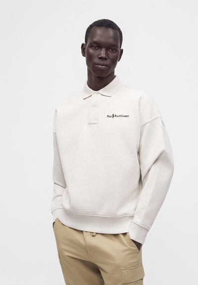 Άνδρας μοντέλο που φοράει λευκό φούτερ Polo Ralph Lauren και μπεζ cargo παντελόνια, στέκεται με τα χέρια στις τσέπες μπροστά σε απλό φόντο.