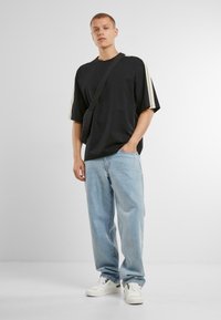 Černé oversized tričko s bílými pruhy na rukávech, světlé modré volné džíny a bílé tenisky, nošené s malou černou crossbody taškou.