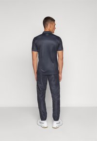 Polo shirt atlético de color navy con mangas cortas, textura suave, combinado con pantalones tapered a juego que presentan una franja negra brillante, y zapatos blancos.