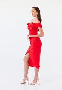 Robe rouge à épaules dénudées avec un corsage ajusté, un motif floral texturé et une fente jusqu'à la cuisse. Associée à des sandales à talons hauts blanches.