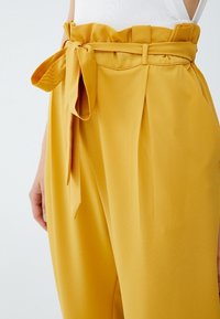 Gula högmidjade byxor i slätt tyg, med en knuten rosett i midjan och veck, samt en vid byxdesign.