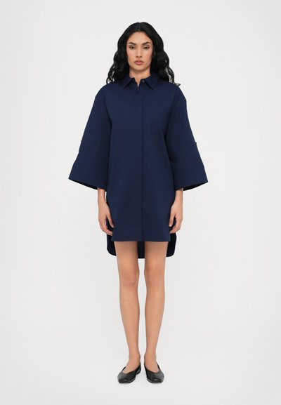 Robe chemise en coton bleu marine avec un col pointu, des manches courtes, une poche avant et un ourlet arrière légèrement plus long. Chaussures plates noires portées.