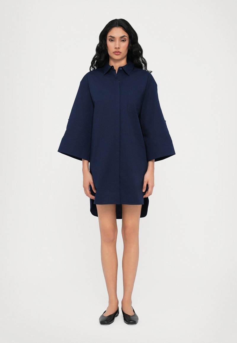 Abito camicia in cotone blu navy con colletto a punta, maniche corte, tasca frontale e orlo posteriore leggermente più lungo. Indossate scarpe nere basse.