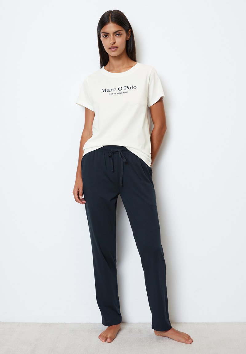 Helles cremfarbige T-Shirt mit schwarzem Text, kombiniert mit dunkelblauen Jogginghosen mit Kordelzug. Beide Artikel haben eine lockere Passform und eine weiche Textur.