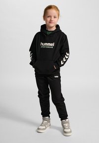 Sudadera con capucha negra con el logo "hummel SPORTSWEAR" y patrón de chevrones blancos en las mangas; combinada con pantalones negros y zapatillas altas grises.