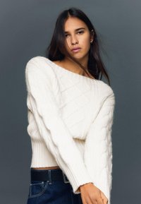 Kr�ém színű, kötött kábelmintás pulóver széles nyakkivágással, bordázott mandzsettákkal és szegéllyel. Sötét farmernadrág fölött viselve, laza szabást mutatva.