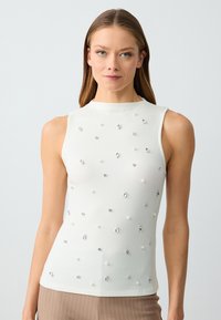 Top senza maniche bianco con un collo alto, adornato da perle e decorazioni metalliche sul davanti, texture liscia.
