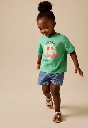 Mladá dívka s kudrnatými vlasy v zeleném tričku s grafikou želé, džínových šortkách a zlatých sandálech, kráčející s mírným úsměvem.