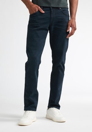 Man die donkerblauwe slim-fit broek en witte sneakers draagt, staat met één hand in de zak tegen een vlakke achtergrond.