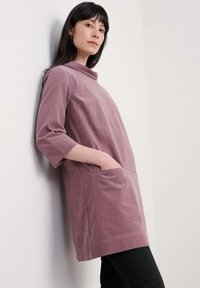 Donna in un vestito tunica di pantalone a maniche lunghe color mauve con tasche, appoggiata a una parete bianca, indossando pantaloni neri.