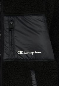 Giacca in pile nera con tasca con zip, caratterizzata da un pannello in nylon liscio e logo "Champion" bianco sul davanti.