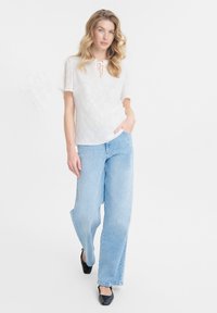 Blusa blanca texturizada con cuello atado, combinada con jeans de pierna ancha de un azul claro y zapatos planos negros. El fondo es neutral.