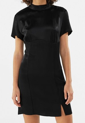 Žena v krátkých rukávech, černých saténových šatech s vysokým výstřihem a předním rozparkem nad kolenem, stojící na jednoduchém pozadí.